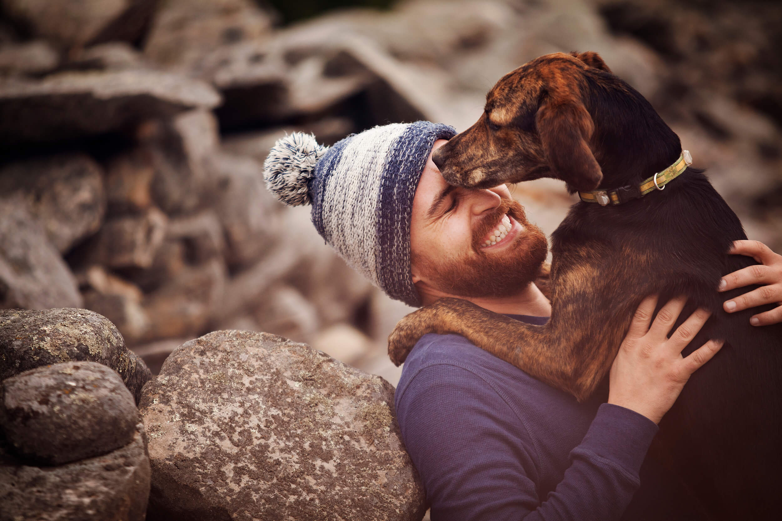 Man with ginger beard and a bobble hat sat down whilst his dog is in his arms. The man is smiling and hugging his dog.