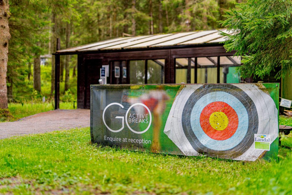 Photograph of the target range at Kielder Waterside Park. The shed is surrounded by trees and features a banner in front with a archery target displayed.