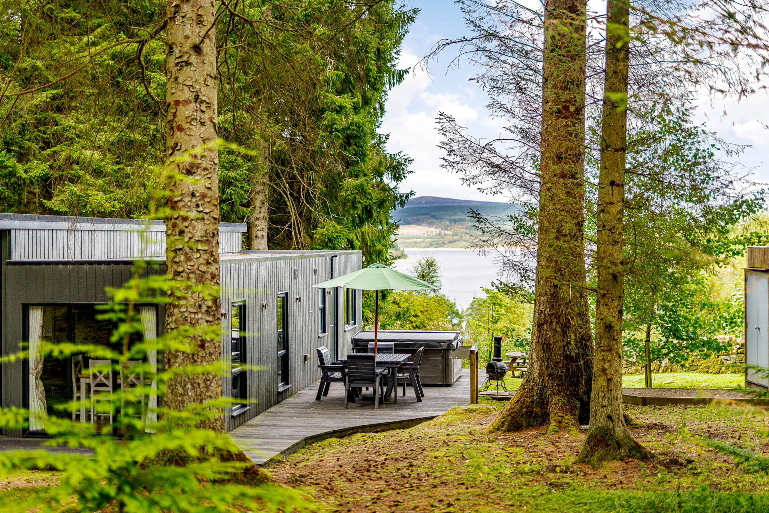 View over the reservoir of a magnificent lodge at Kielder Waterside Park. The lodge looks modern and features a hot tub outside, surrounded by tall trees and green grass.